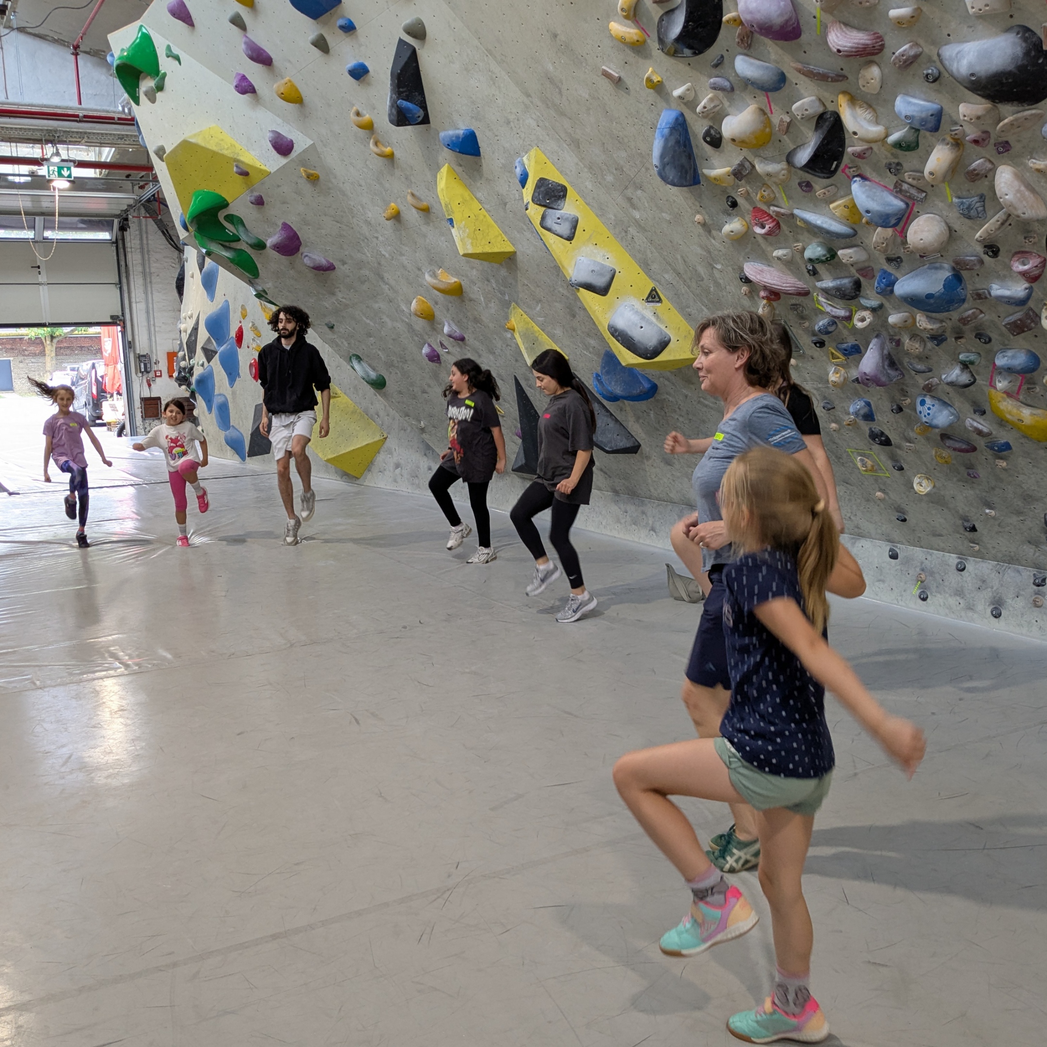 Kinder springen im Halbkreis als Aufwärmübung vor einer Kletterwand.