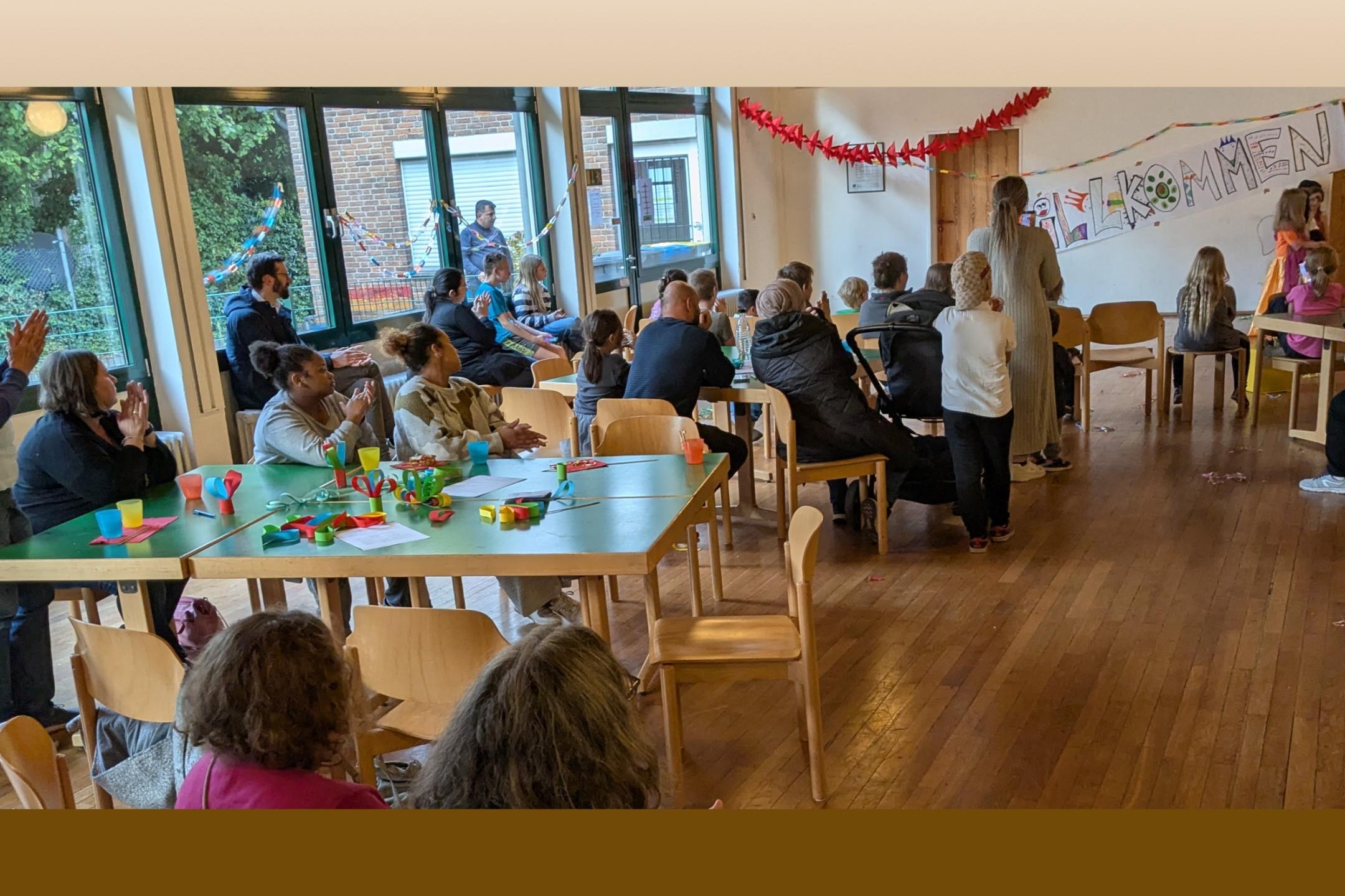 Menschen unterschiedlichen Alters sitzen an Tischen und schauen nach vorne zu einem Willkommens-Schild.