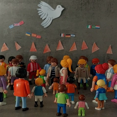 Viele Playmobilmännchen stehen zusammen. Über ihnen Feuerzungen und am Himmel eine weiße Taube.