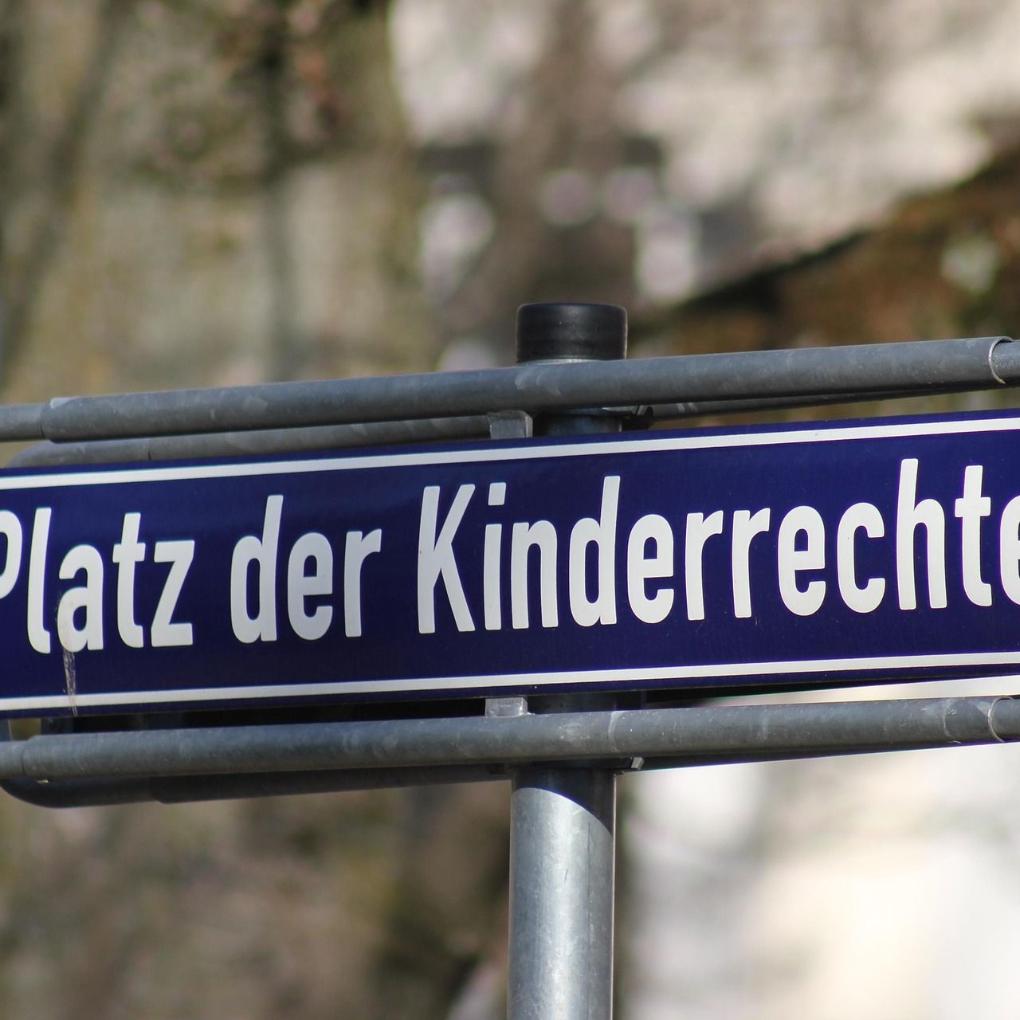 Auf einem blauen Schild steht in weißer Schrift: „Platz der Kinderrechte“. Das Schild hängt in einem grauen Metall-Rahmen. Im Hintergrund sind Bäume und Häuser zu erkennen. Der Hintergrund ist unscharf.