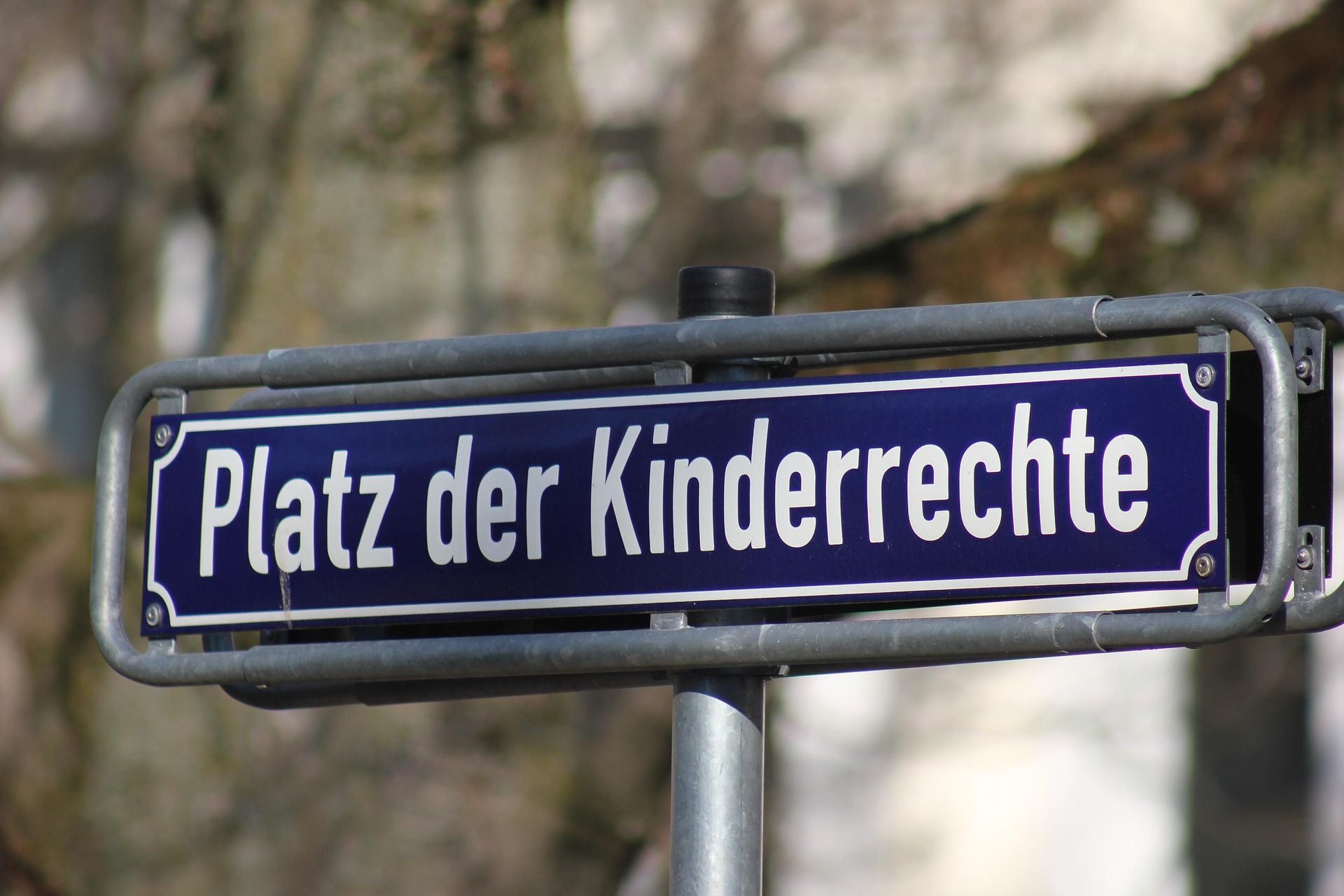Auf einem blauen Schild steht in weißer Schrift: „Platz der Kinderrechte“. Das Schild hängt in einem grauen Metall-Rahmen. Im Hintergrund sind Bäume und Häuser zu erkennen. Der Hintergrund ist unscharf.