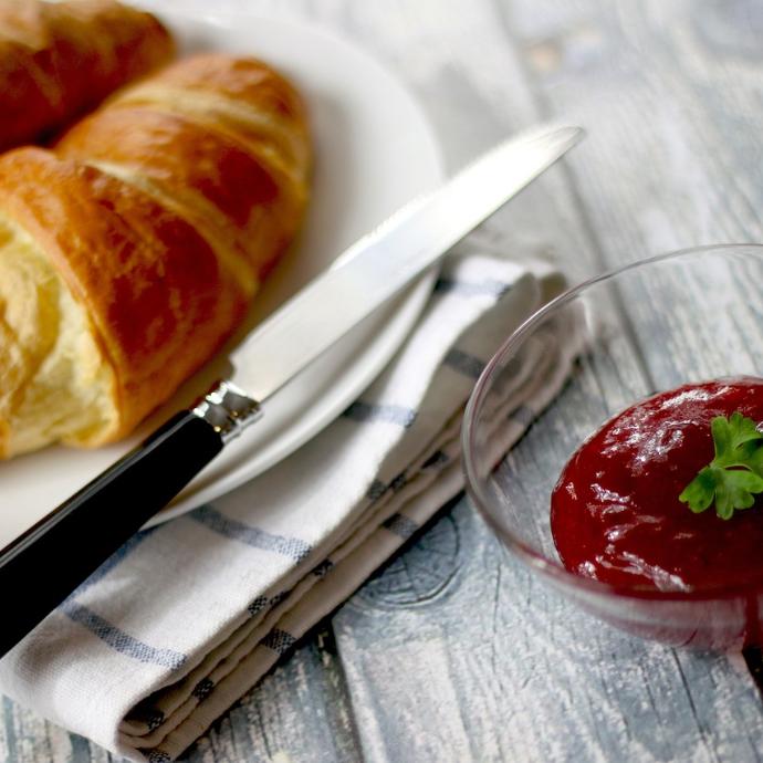Frühstücksteller mit 2 Croissants, einem Messer und einer Serviette abgebildet. Daneben steht ein Schälchen roter Marmelade.