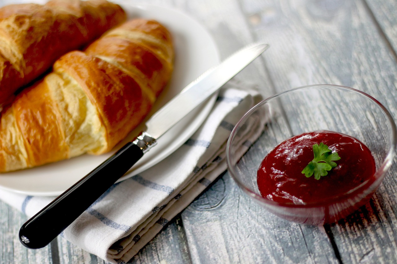 Frühstücksteller mit 2 Croissants, einem Messer und einer Serviette abgebildet. Daneben steht ein Schälchen roter Marmelade.