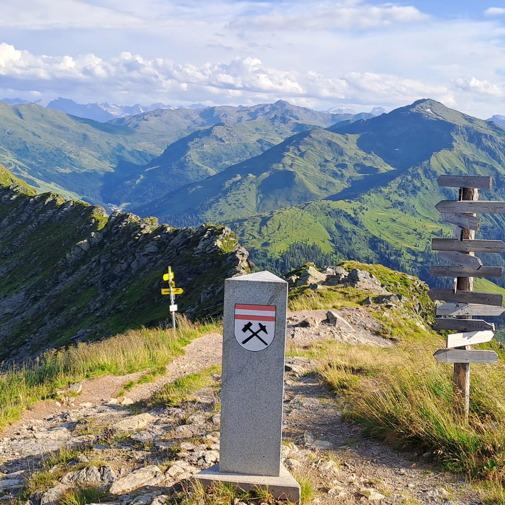 Ein schmaler Wanderweg führt über einen grünen Bergkamm. In der Mitte steht ein Grenzstein mit Symbol. Daneben stehen mehrere Wegweiser. Im Hintergrund sind weite Berge und Täler zu sehen.