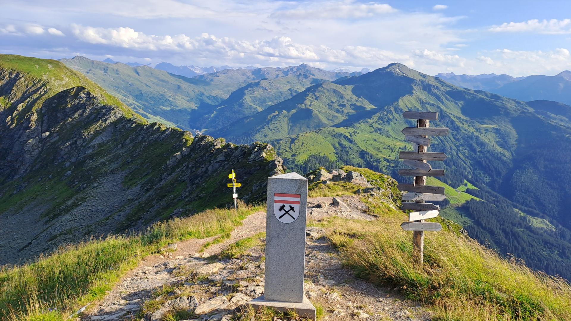 Ein schmaler Wanderweg führt über einen grünen Bergkamm. In der Mitte steht ein Grenzstein mit Symbol. Daneben stehen mehrere Wegweiser. Im Hintergrund sind weite Berge und Täler zu sehen.