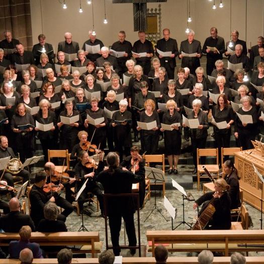 Viele Chormitglieder singen in Formation, davor steht der Dirigent mit Orchester.