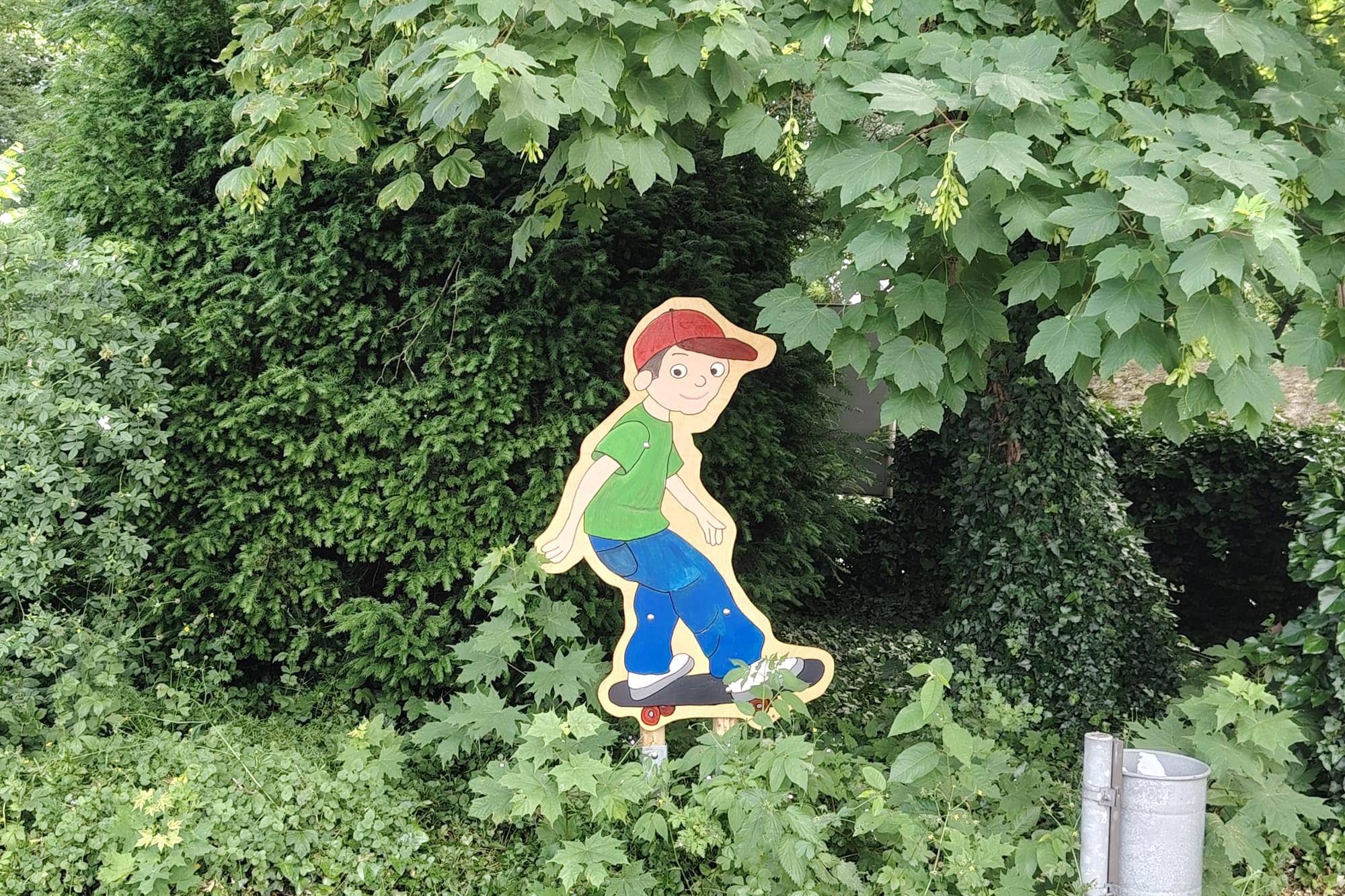 Im Grünen ist ein Junge auf einem Skateboard mit einer roten Käppi, grünem T-Shirt und einer blauen Hose.